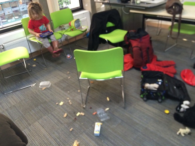 Part of a university meeting room, showing a toddler playing on a tablet, sitting on a chair. The floor is covered in dropped food and strewn toys.